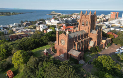 Aerial view of Christ Church Cathedral in Newcastle, Australia, surrounded by lush green trees and urban buildings, with the coastline and ocean visible in the background under a clear blue sky.