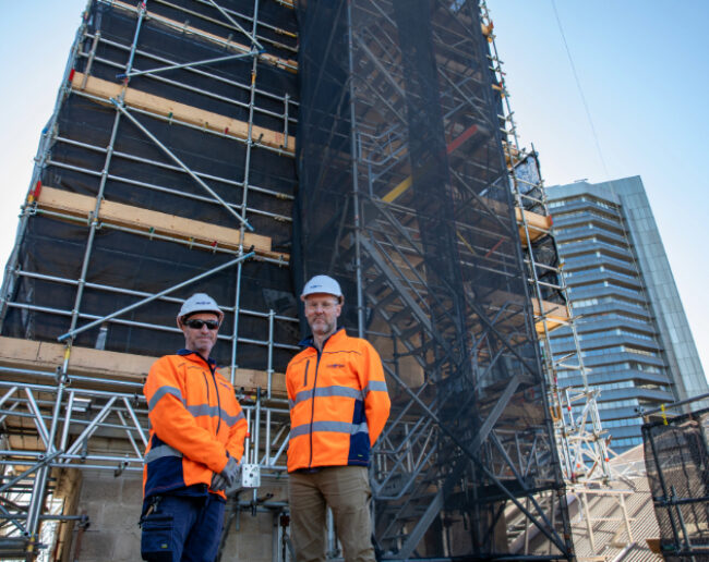 Two men in orange jackets and hard hats in front of scaffolding